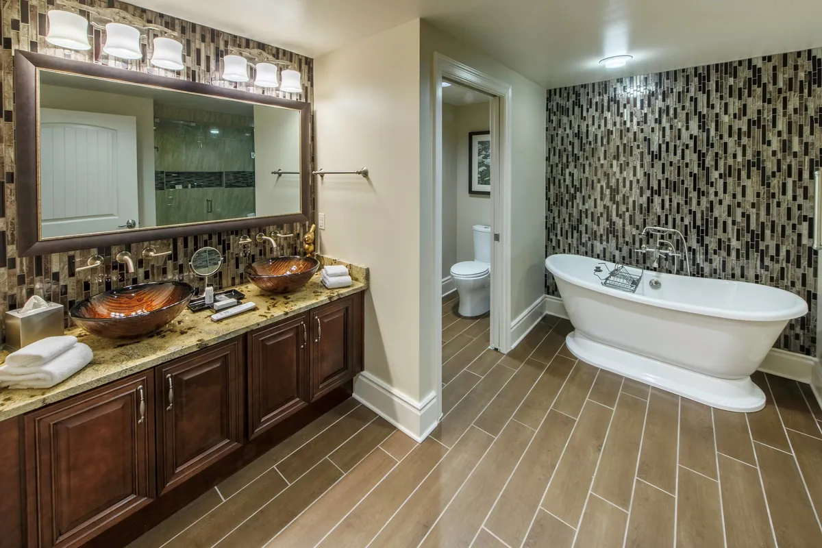 An upscale, tranquil bathroom with double vanity sinks, a freestanding spa bathtub, and a decorative mosaic accent wall. The scene invites relaxation and self-care, echoing the spa-like features described in the master suite.