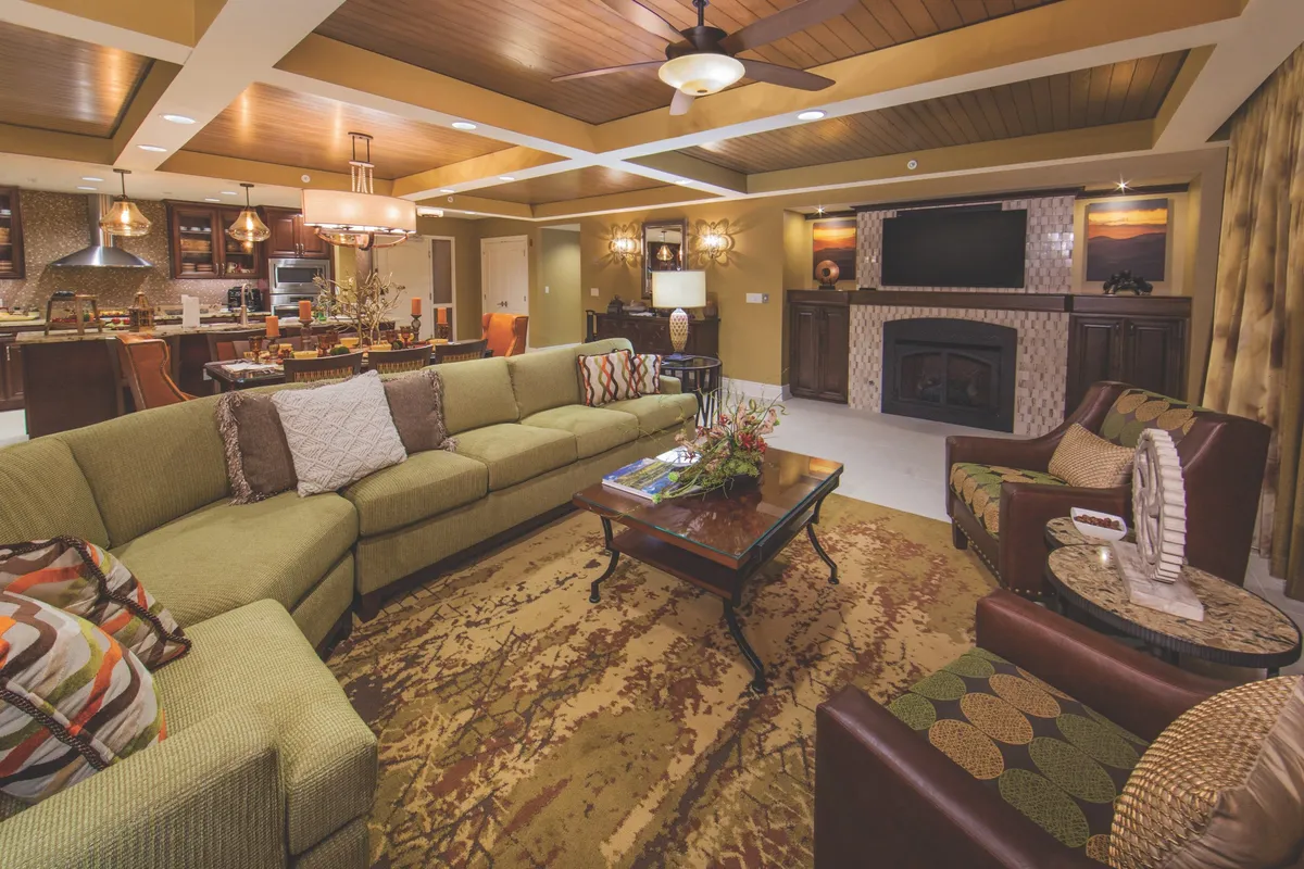 A spacious living room featuring a green sectional sofa centered around a wooden coffee table, with a fireplace creating a cozy focal point. The room is brightened by natural light streaming through large windows, and a dining area is visible in the background, suggesting a seamless flow for gatherings and shared meals.