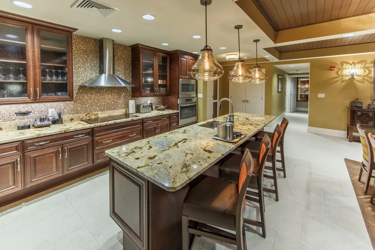 This image features a warm and inviting kitchen with granite countertops, wood cabinetry, hanging pendant lights, and modern stainless steel appliances. The space is clearly designed for cooking and socializing, with ample surface area for food prep and plenty of room for multiple people to gather. The mood is cozy and communal, suggesting both lively family feasts and quiet everyday meals.