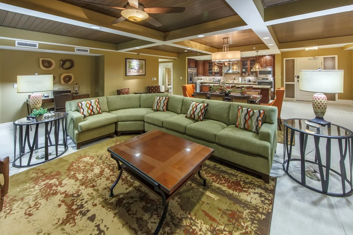 The image displays a warm, inviting living area with a spacious curved sectional sofa and elegant wooden coffee table, complemented by tasteful art on the walls. In the background, a modern kitchen invites guests to gather, while the overall ambiance suggests a perfect space for socializing, relaxing, and making memories with loved ones.