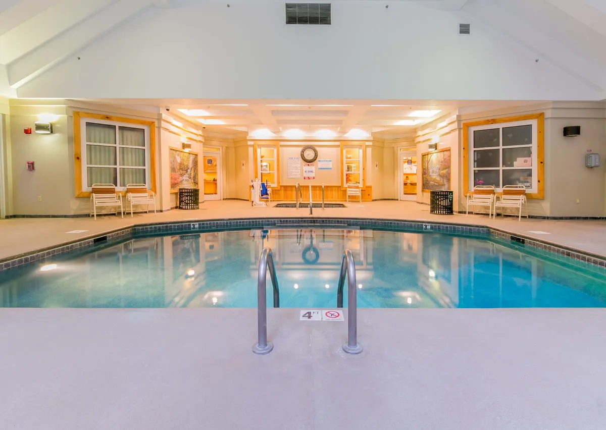 A serene indoor pool area with a sparkling swimming pool, stainless steel ladder, comfortable seating, and a bright reception space. Sunlight streams in, creating a tranquil atmosphere ideal for morning swims or evening relaxation.