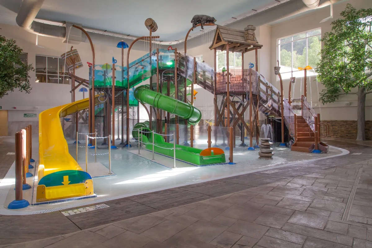 A vibrant indoor water park scene filled with colorful water slides, a play structure, and interactive water features, surrounded by lush faux trees. Children and families are actively playing and enjoying various water activities in a joyful, lively setting.