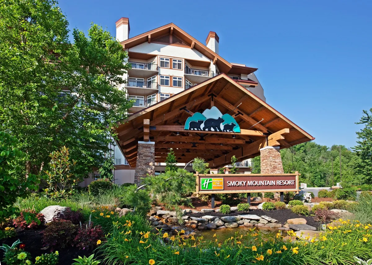 This exterior image showcases the resort's entrance with a welcoming canopy, a clearly visible resort sign, and landscaped gardens set against the backdrop of the surrounding mountains. The building facade is inviting, and the scene alludes to the property's close integration with the natural environment of the Smokies.