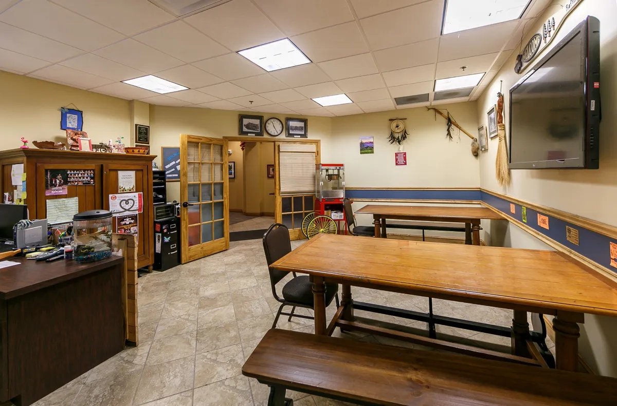 This image presents a friendly and welcoming dining area with wooden tables, a wooden desk, television, and decorative wall touches. The setup encourages group dining, shared meals, and conversation, evoking the feeling of communal feasts and relaxed family evenings.